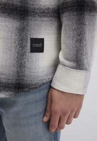 Black and white plaid flannel shirt with a soft texture, featuring a Mavi logo patch. Long sleeves show cuff detail against denim.