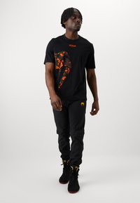 Sort T-shirt med korte ærmer i orange og sort camouflage-design, parret med sorte slim-fit bukser og sorte høj-top sneakers med guld accenter.