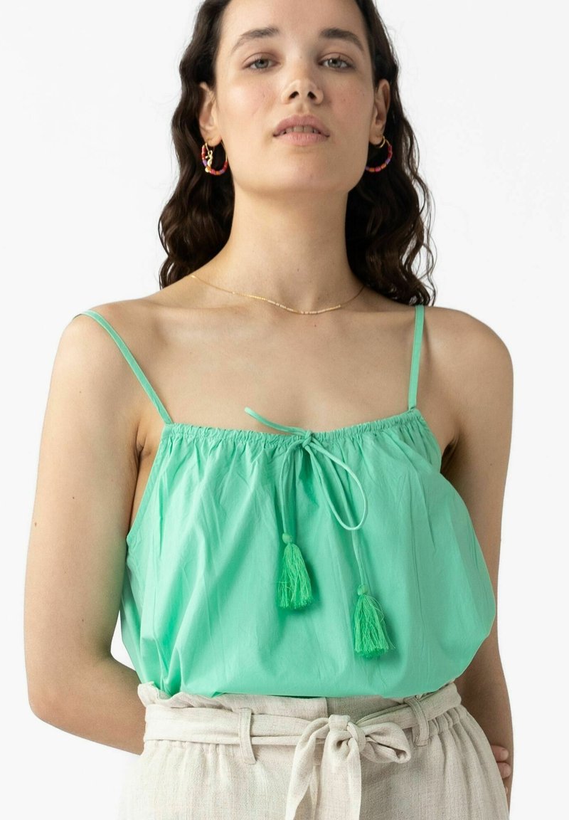 Top sans manches vert menthe en tissu léger, présentant un col froncé avec un nœud et deux pompons verts en accent.