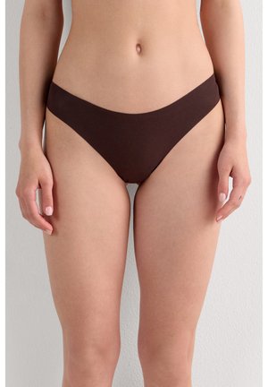 Kobieta w bezszwowej, ciemnobrązowej bieliźnie bikini o wysokim kroju stojąca na tle jednolitej jasnej ściany.