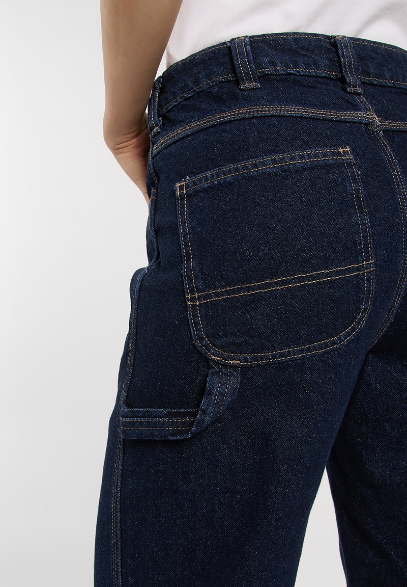 Person wearing dark blue denim jeans with visible back and side pockets, standing against a plain white background.