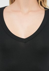 Μαύρο τοπ με λαιμόκοψη σε σχήμα V, κατασκευασμένο από λείο και ελαστικό ύφασμα. Διαθέτει εφαρμοστή γραμμή και καθαρό φινίρισμα γύρω από τον λαιμό. Χωρίς ορατά σχέδια ή διακοσμητικά στοιχεία.