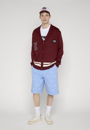 Młody mężczyzna ubrany w bordowy kardigan na guziki, białą koszulkę, fioletowe szorty z białym napisem, białe skarpety, buty sportowe oraz dopasowaną czapkę.
