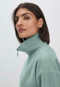 Pull léger en polaire zippé de couleur vert menthe, doté d'un col haut, d'une texture douce et d'une coupe décontractée, avec des coutures accentuées sur les épaules.