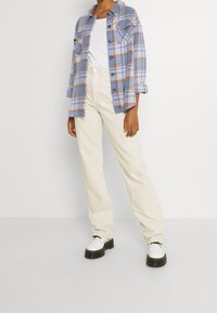 Persona vestita con pantaloni beige, camicia bianca, camicia a quadri blu e marrone con bottoni e scarpe bianche con suola spessa, in piedi contro un muro bianco.