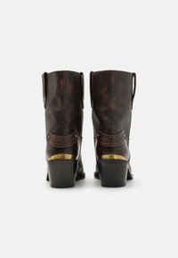 Bottes chelsea en cuir marron avec un talon empilé, présentant des coutures visibles, un branding décoratif, et un accent en laiton distinctif sur le talon.