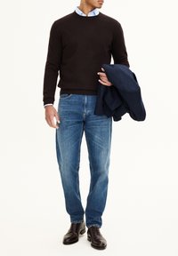 Brauner Pullover, hellblaues Hemd mit Kragen, blaue Jeans und dunkelbraune Lederschuhe. Marineblauer Blazer über dem linken Arm getragen.