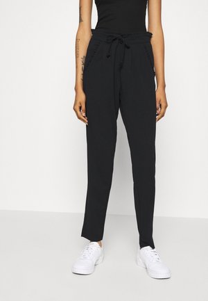 Femme portant un pantalon noir taille haute style paper bag avec une ceinture nouée et des baskets blanches, debout devant un fond blanc uni.