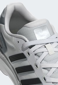 Zapatilla deportiva blanca con parte superior de malla, acentos de goma y cordones a tono. Incluye tres rayas negras y un parche de logo metálico.
