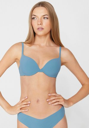 Woman with long blonde hair wearing matching blue bra and underwear, standing with hands on hips, looking to the side.
