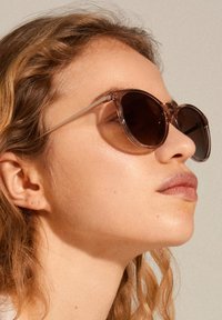 Round, transparent brown sunglasses with dark lenses; thin, clear temples; features a minimalistic and modern design with glossy finish.