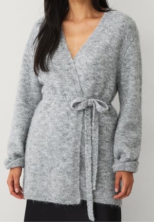 Femme portant un pull-over doux, gris clair, manches longues, noué à la taille, sur une jupe noire sur fond uni.