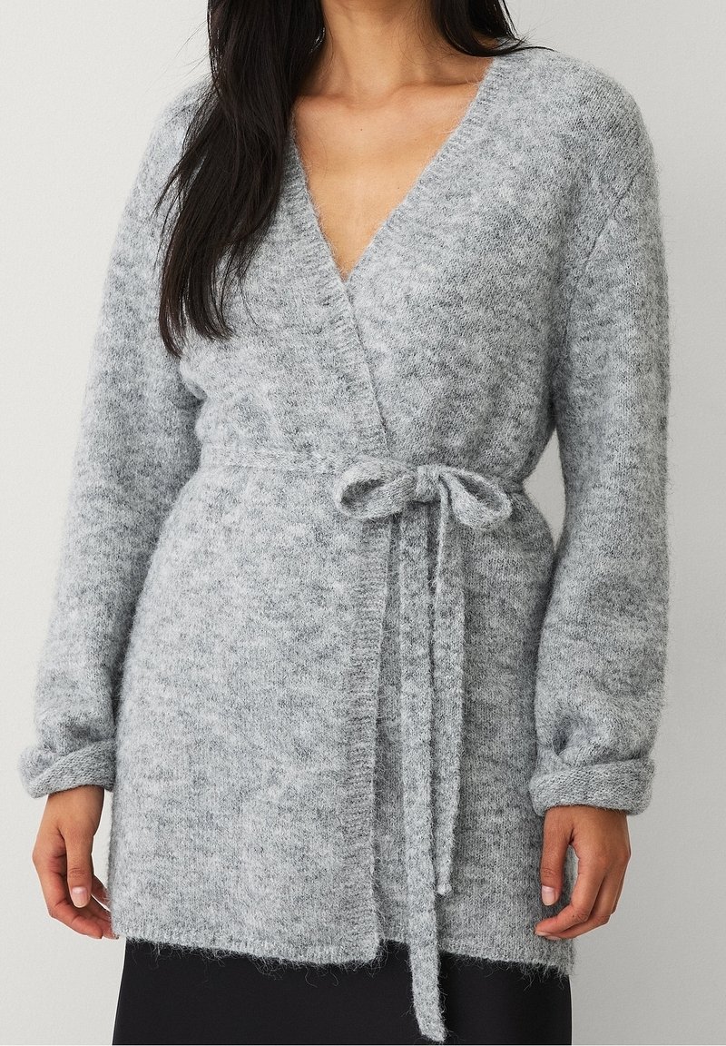 Femme portant un pull-over doux, gris clair, manches longues, noué à la taille, sur une jupe noire sur fond uni.