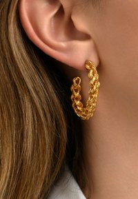 Boucle d'oreille en or avec un design tressé et des détails texturés, présentée sur un fond de tons de cheveux et de peau.