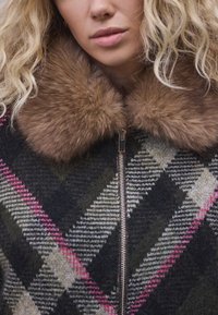 Strickjacke mit geometrischem Muster in Dunkelgrün, Schwarz und Rosa; mit braunem Pelzkragen und silbernem Reißverschluss.