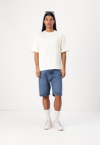 Kremfarget t-skjorte med korte ermer og en subtil logo, kombinert med blå denimshorts, hvite sneakers og hvite sokker i ankellengde.