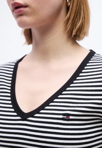 Γυναίκα με κοντά ανοιχτό καστανά μαλλιά, φορώντας μαύρο και λευκό ριγέ μπλουζάκι με V λαιμόκοψη και μικρό κόκκινο, λευκό και μπλε λογότυπο στο στήθος.