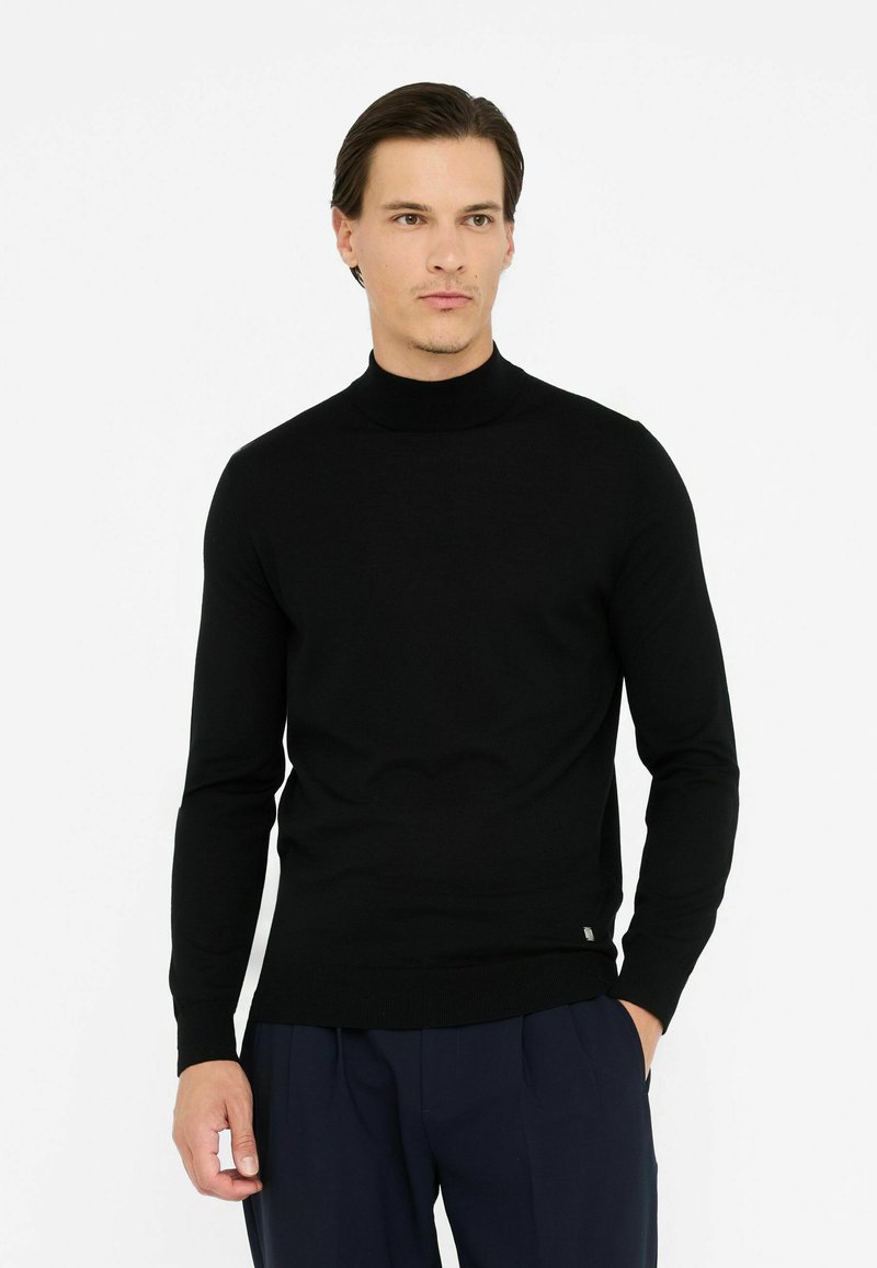 Pull à col roulé noir à manches longues, texture tricot douce, coupe ajustée, avec un petit logo en détail sur l'ourlet. Associé à un pantalon sombre.