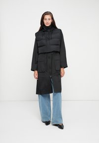 Zwarte gewatteerde vest gelaagd over een lange zwarte jas met een getailleerde taille, gecombineerd met lichtblauwe wijde jeans en zwarte schoenen.