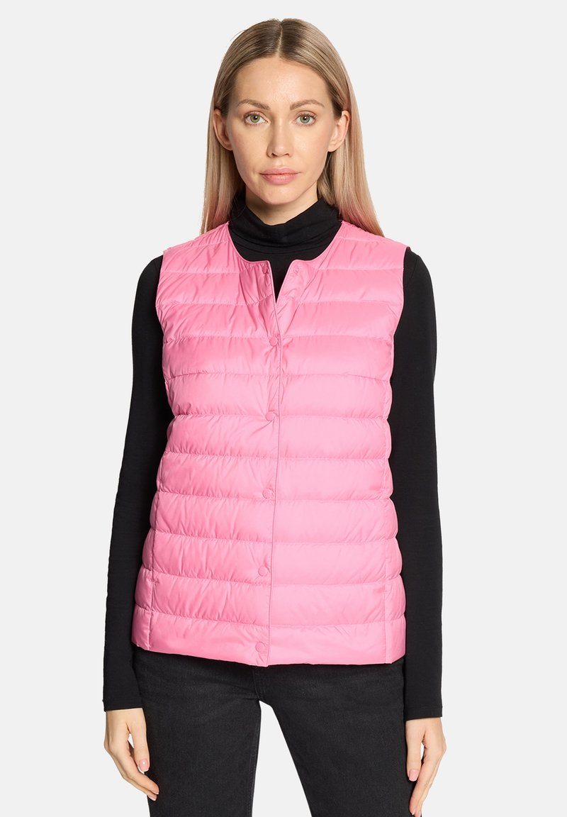 Donna che indossa un gilet trapuntato rosa senza maniche sopra una maglia nera a maniche lunghe e pantaloni neri, in piedi davanti a uno sfondo chiaro e uniforme.