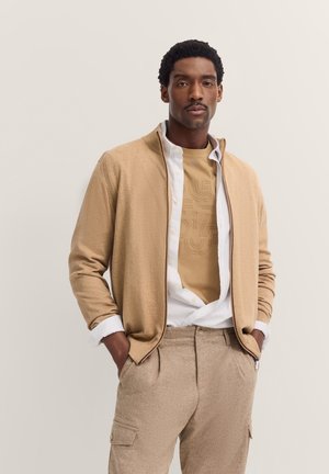 Beige Strickjacke mit Reißverschlusskragen, übereinander getragen mit einem hellbraunen Grafik-T-Shirt, kombiniert mit hellbraunen, strukturierten Cargo-Hosen. Lässiges Ensemble.