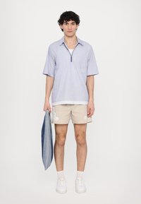 Camicia azzurra leggera con maniche corte e cerniera, dotata di taschino; pantaloni corti beige con ricamo floreale bianco; in mano una giacca di jeans; sneakers bianche.