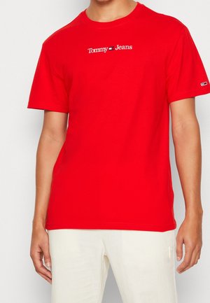 T-shirt rouge à manches courtes avec le logo "Tommy Jeans" en blanc et bleu marine sur la poitrine, porté avec un pantalon blanc cassé.