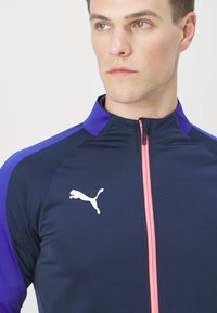 Temno modra športna jakna s purpurnimi detajli, z zadrgo spredaj in belim logotipom Puma na prsih, s teksturiranim dizajnom.