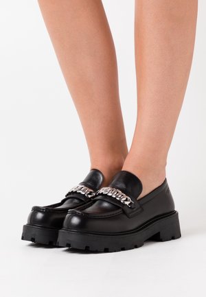 Černé loafers s robustní podrážkou a stříbrným řetízkovým detailem, nošené na bosých nohou na bílém pozadí.