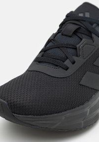Gros plan d'une chaussure de sport noire avec tige en mesh, languette rembourrée, lacets noirs et semelle texturée sur fond blanc.