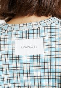 Niebiesko-biała tkanina w kratę z czarnymi akcentami, z prostokątną białą etykietą z napisem „Calvin Klein” w czarnym fontcie z tyłu.