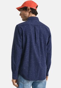 Marineblauwe overhemd met lange mouwen en een gespikkeld patroon, knopmanchetten en een rugplooi, gecombineerd met lichte denim jeans en een rode pet.