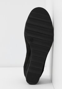 Semelle d’une chaussure noire avec une semelle texturée et un marquage de taille, positionnée à la verticale contre un fond blanc angulaire.
