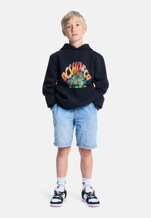 Hoodie noire avec logo graphique coloré, short en jean clair et baskets noires avec accents blancs et marque rouge. Chaussettes blanches visibles.