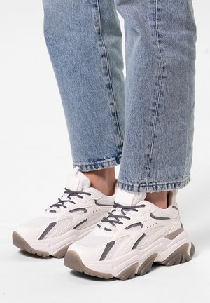 Chaussures : baskets blanches épaisses avec des accents gris, en matériaux en mesh et synthétiques, semelles texturées et talons en caoutchouc épais. Portées avec un jean bleu retroussé.