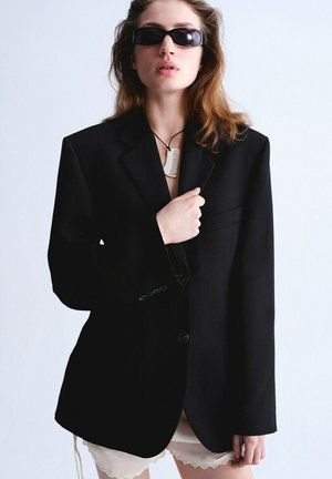 Mujer con blazer negro de gran tamaño, gafas de sol estrechas negras y un collar colgante en forma de peine blanco, posando contra un fondo liso.