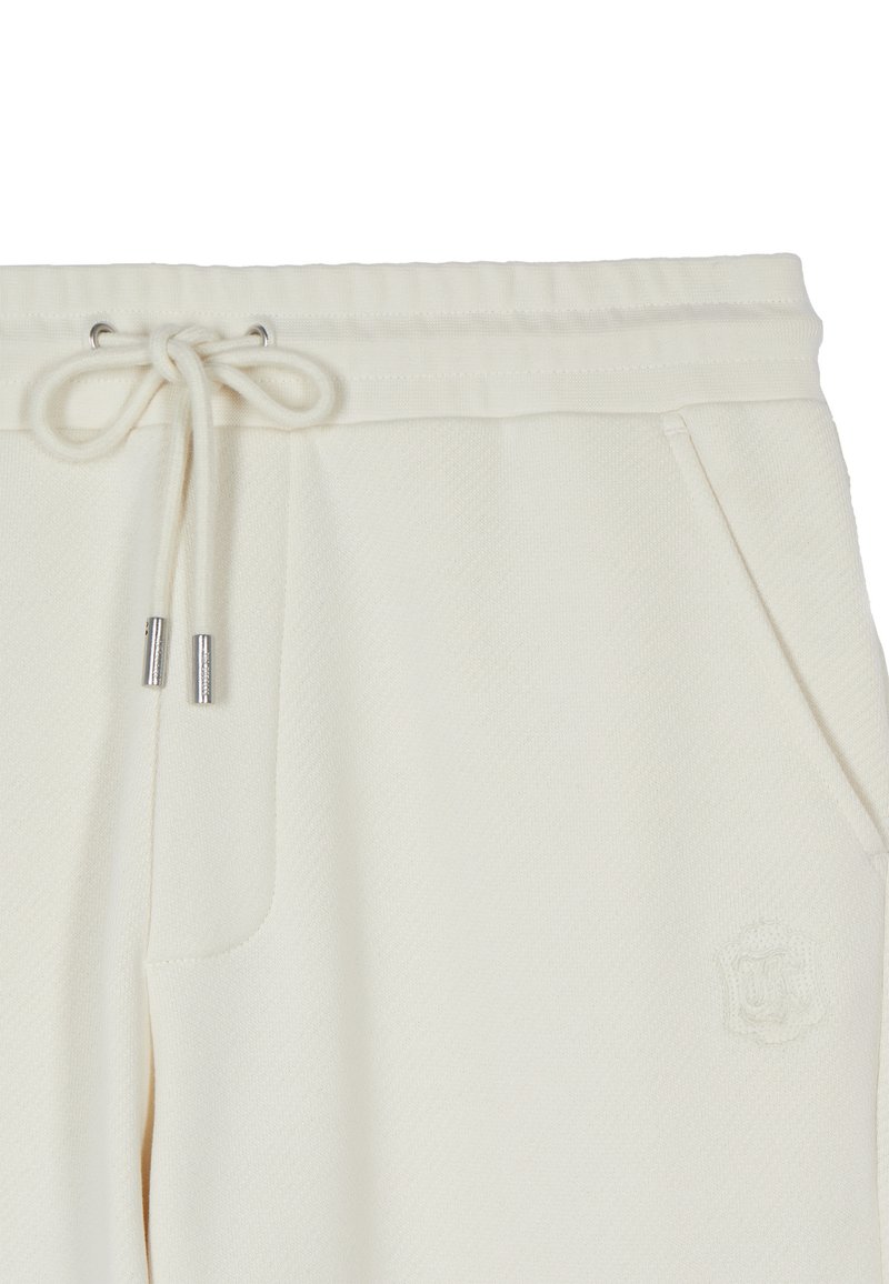 Primer plano de pantalones deportivos color crema con cintura de cordón, cordones con punta metálica, bolsillos delanteros y logo bordado sutil en el lado derecho.