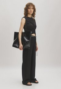 Schwarze Tragetasche mit glänzender Oberfläche und minimalistischem Design, kombiniert mit einem taillierten schwarzen Oberteil und weiten schwarzen Hosen. Glatte Textur, elegante Form.