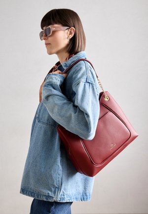 Sac en cuir rouge avec une surface texturée, dotée de quincaillerie dorée et d'un accent chaîne. Porté sur l'épaule contre une veste en denim.