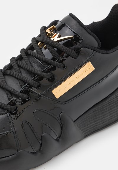 Sneaker noir avec tige en cuir verni, accents texturés, plaque de marque dorée et semelle épaisse avec des éléments de design crantés.