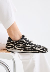 Zapatillas de deporte con estampado de cebra en negro y beige, con cierres de cordones, parte superior texturizada y suela negra, llevadas con pantalones blancos sobre una silla.