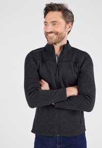 Chaqueta de forro polar negra con cremallera, textura suave, cuello alto y detalle sutil del logo en el hombro. Llevada sobre una camiseta gris clara.