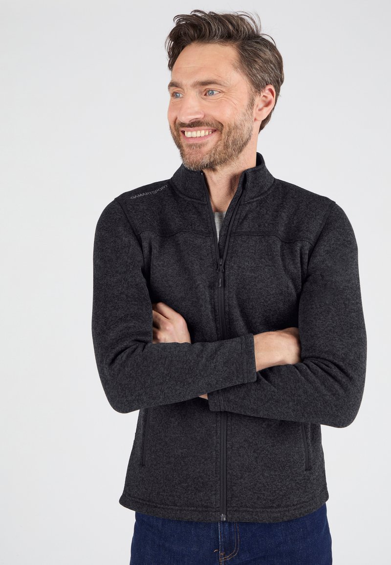 Chaqueta de forro polar negra con cremallera, textura suave, cuello alto y detalle sutil del logo en el hombro. Llevada sobre una camiseta gris clara.
