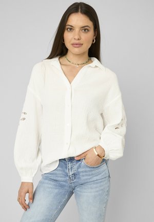 Femme aux longs cheveux foncés portant un chemisier blanc texturé avec des manches découpées et un jean bleu clair, accessoirisée avec des bijoux.