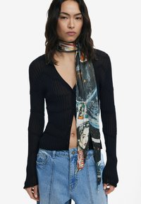 Schwarze gerippte Strickjacke mit langen Ärmeln, kombiniert mit einem mehrfarbigen gemusterten Schal mit Illustrationen. Jeans aus Denim sind am Taillenbereich sichtbar.