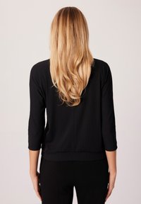 Czarna bluzka z trzema ćwiartkami rękawów o prążkowanej fakturze, luźny krój i prosty dół. Włosy są długie i faliste, spływające w dół pleców.