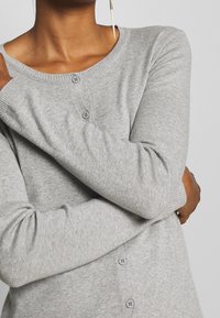 Szary, dziany kardigan z okrągłym dekoltem, zapięciem na guziki z przodu i długimi rękawami. Posiada miękką teksturę i ściągacze przy mankietach.