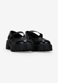 Zapatos de plataforma negros con puntera redondeada, correa en el empeine y suela de goma texturizada con profundas estrías. Parte superior similar al cuero suave.