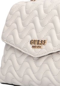 Sac à main crème matelassé avec une texture ondulée, arborant un logo doré "GUESS" et un accent "EST 1981". Fermeture à rabat magnétique.