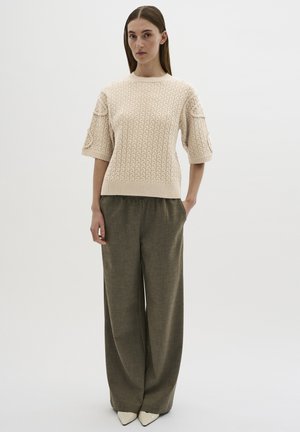 Beiger Strickpullover mit kurzen Ärmeln, der ein strukturiertes Muster aufweist; kombiniert mit lockeren grau-grünen Hosen und spitzen weißen Schuhen.